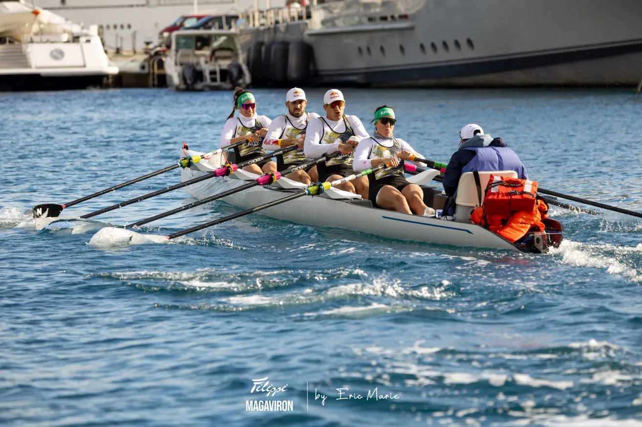Ein Ruderteam in einem Boot mit Red-Bull-Markenzeichen, das auf einem Fluss rudert, mit einem anderen Boot und einem Steg im Hintergrund.