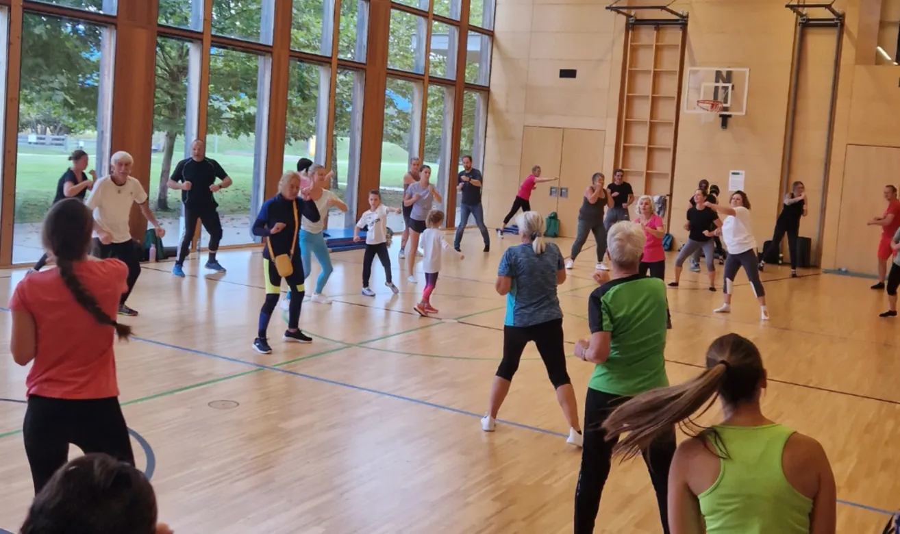 Eine Gruppe von Menschen trainiert in einem Fitnessstudio. Eine Person vorne leitet das Training. Rechts befindet sich ein Basketballkorb.