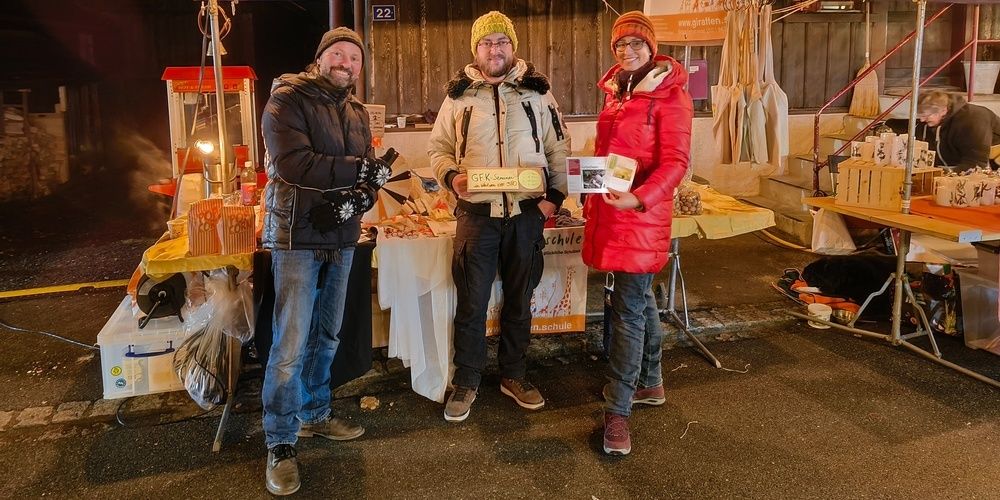 Drei Personen stehen lächelnd in einer festlichen Umgebung. Zwei Männer und eine Frau. Ein Tisch hinter ihnen ist mit einem gelben Tuch bedeckt.