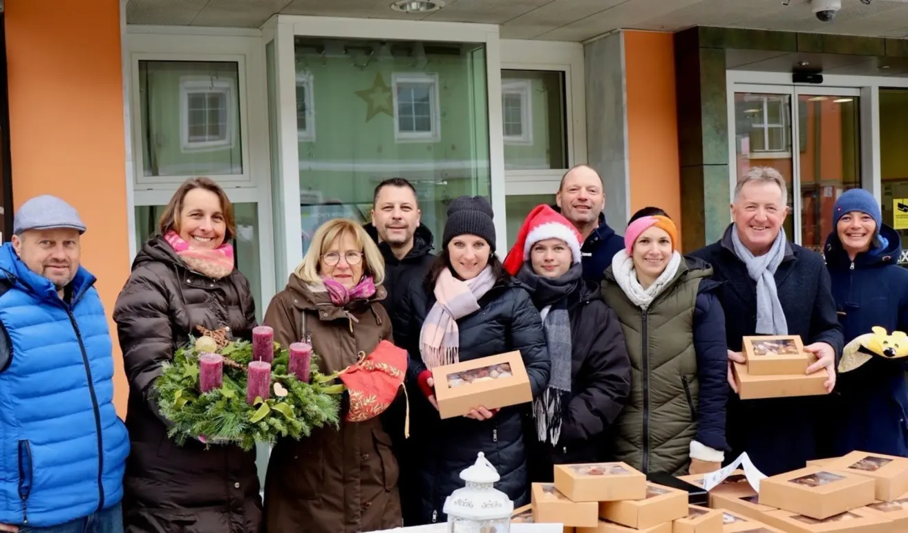 Eine Gruppe von Menschen steht vor einem Gebäude mit einem Kranz und Boxen. Sie alle lächeln und scheinen für ein Foto zu posieren. Sie tragen Winterkleidung.