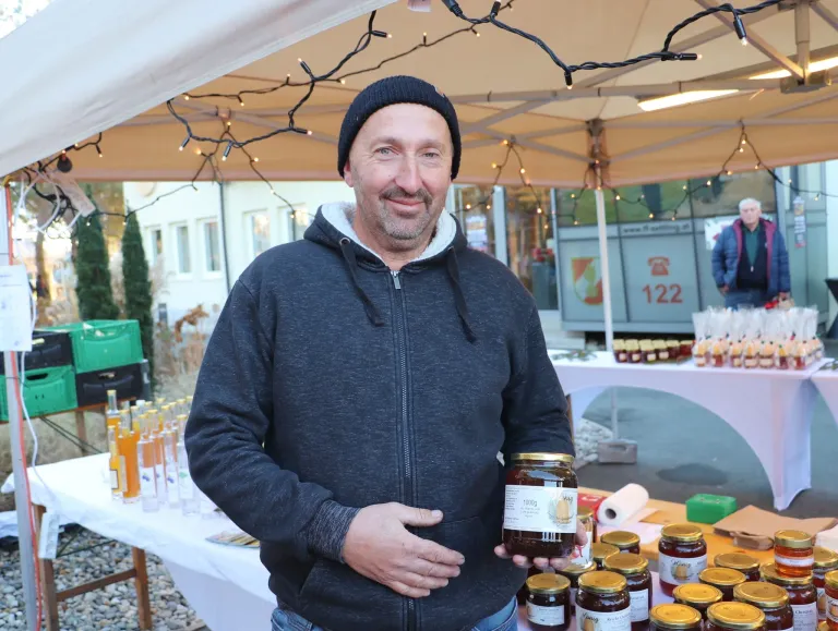 Ein Mann in einer blauen Kapuzenjacke steht an einem Marktstand, lächelt und hält ein Glas Honig in der Hand. Hinter ihm befindet sich ein Baldachin mit Lichterketten, und mehrere Gläser Honig sind auf dem Tisch ausgestellt. Im Hintergrund steht ein Gebäude mit einem Schild, auf dem die Nummer 122 zu sehen ist.
