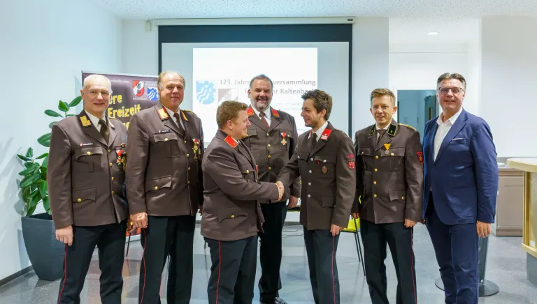 Fünf Männer in Militäruniformen stehen in einer Reihe, zwei schütteln Hände. Sie stehen vor einer Präsentationsleinwand und einem Banner.
