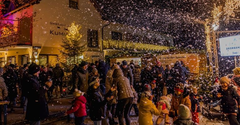 Menschen, Erwachsene und Kinder, genießen eine verschneite Nacht mit fallenden Schneeflocken vor einem Gebäude mit einem Weihnachtsbaum und Lichtern.