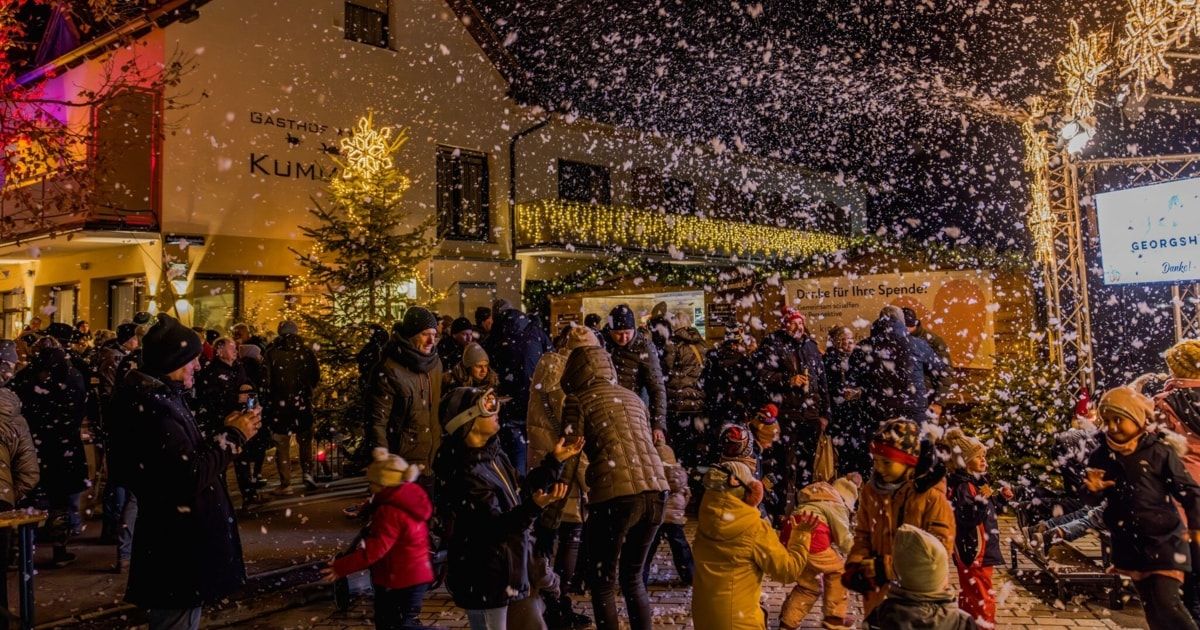 Menschen, Erwachsene und Kinder, genießen eine verschneite Nacht mit fallenden Schneeflocken vor einem Gebäude mit einem Weihnachtsbaum und Lichtern.