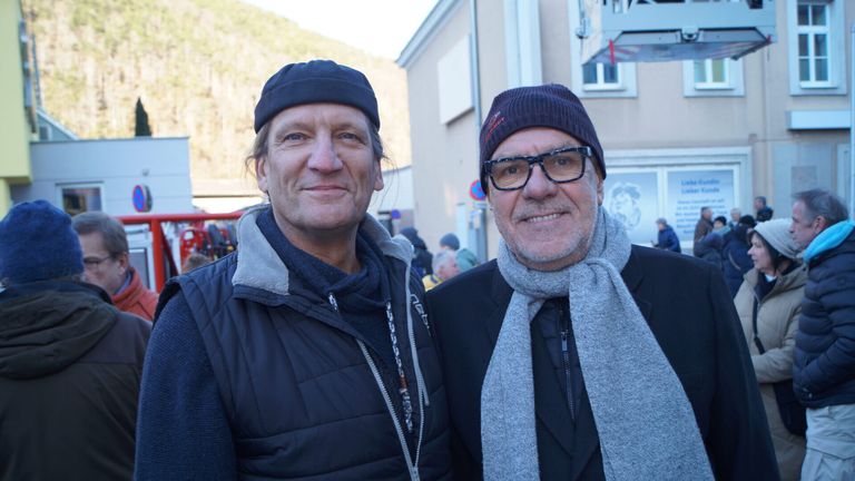 Zwei Männer stehen lächelnd zusammen in einem öffentlichen Bereich mit einem Gebäude und Bergen im Hintergrund. Beide tragen Winterkleidung.