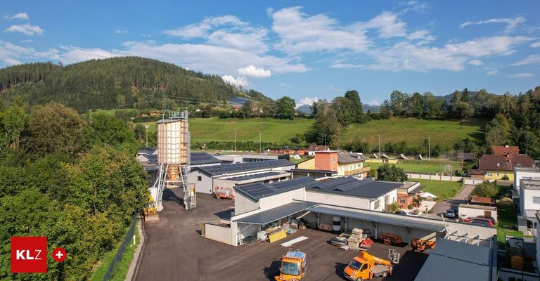 Ein Blick von oben auf eine Industrieanlage mit Solarmodulen auf dem Dach und mehreren Fahrzeugen auf dem Parkplatz. Im Hintergrund befinden sich Hügel, Bäume und Häuser.