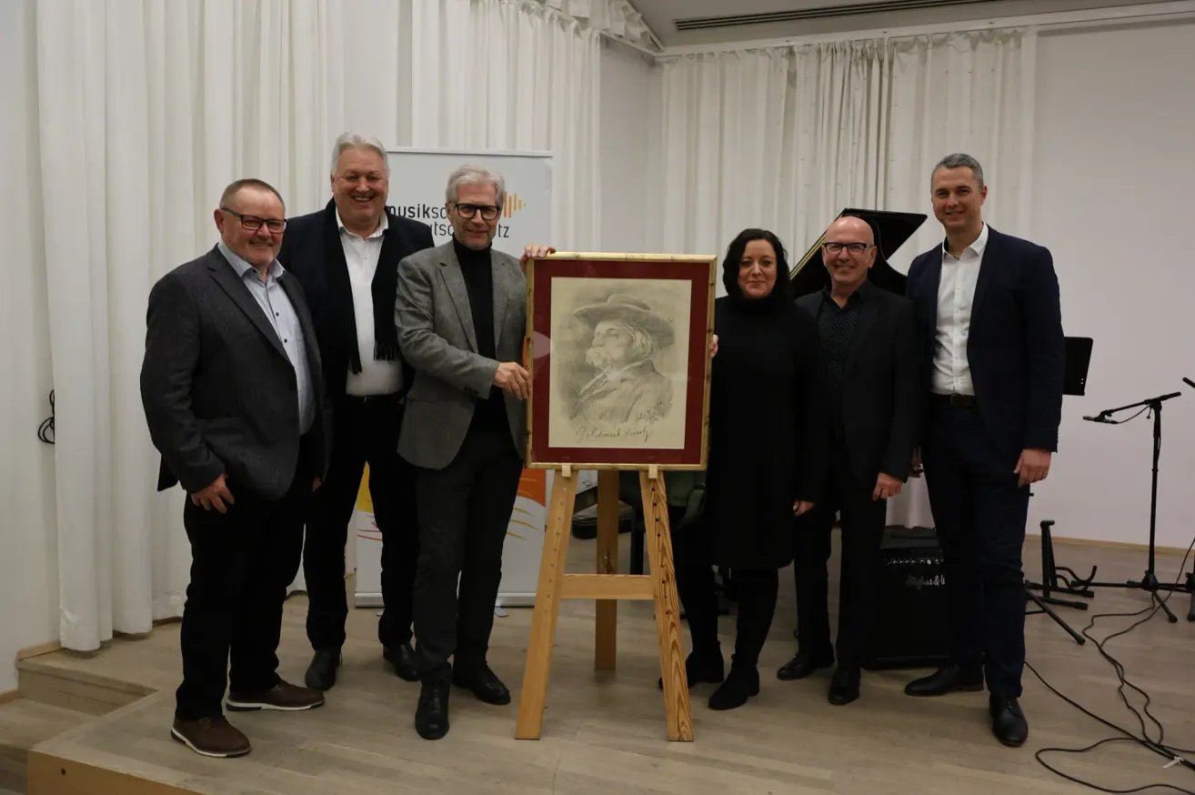 Sechs Personen stehen und posieren für ein Foto mit einem Porträt in einem Holzrahmen. Zwei Männer halten den Rahmen, und ein anderer Mann lächelt. Ein Klavier steht hinter ihnen, und weiße Vorhänge sind an der Wand.