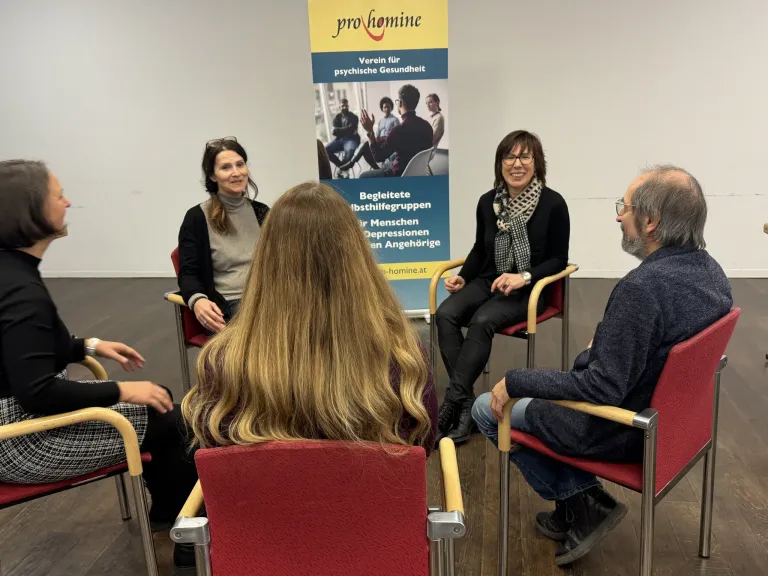 Eine Gruppe von Menschen ist in einem Raum versammelt und führt eine Diskussion. Eine Frau mit einem Schal lächelt und sieht eine andere Frau an. Ein Banner hinter ihnen trägt den Text 'pro hömine'.