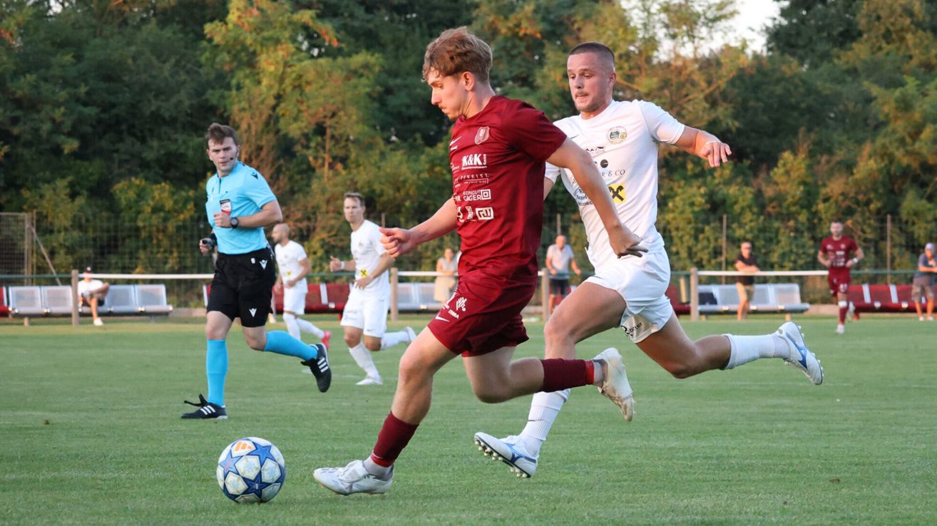 Ein Fußballspiel mit Spielern in burgunderroten und weißen Trikots auf einem Rasenplatz mit einem verschwommenen Hintergrund aus Bäumen und Menschen.