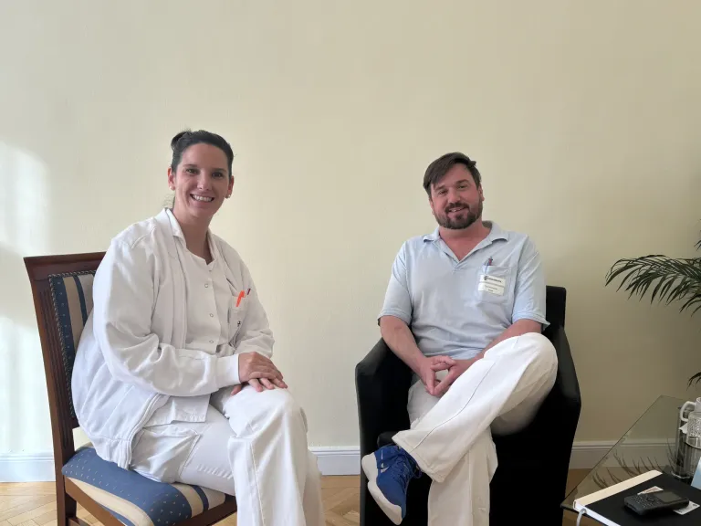 Eine Frau in einem weißen Laborkittel sitzt auf einem Stuhl mit den Händen im Schoß und lächelt. Neben ihr sitzt ein Mann in einem blauen Hemd mit Namensschild auf einem schwarzen Stuhl mit verschränkten Beinen. Sie befinden sich in einem Raum mit einer beigen Wand.