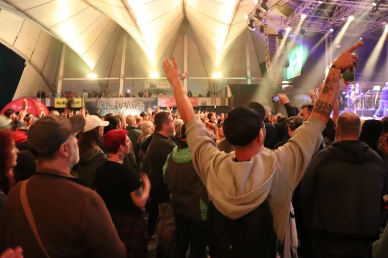 Eine große Menschenmenge ist in einem kuppelförmigen Veranstaltungsort versammelt. Viele Menschen heben ihre Hände, und einige machen Fotos. Die Bühne hat Banner und eine grüne Leinwand. Scheinwerfer sind auf die Bühne gerichtet, und die Decke hat Metallstrukturen.