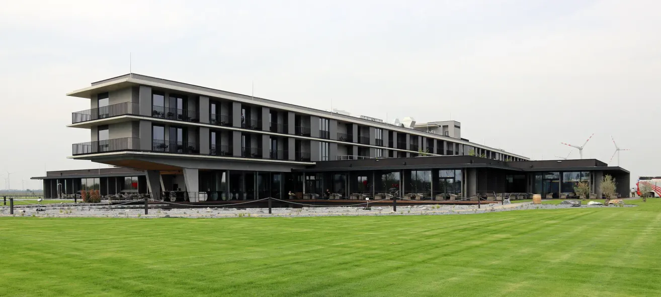 Ein modernes Hotel mit mehreren Stockwerken und Glasfenstern steht auf einem grünen Rasen. Einige Leute sind auf der Terrasse, und mehrere Autos sind davor geparkt.