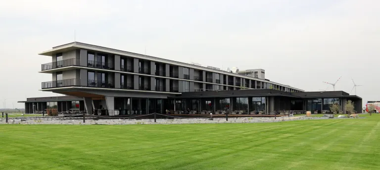 Ein modernes Hotel mit mehreren Stockwerken und Glasfenstern steht auf einem grünen Rasen. Einige Leute sind auf der Terrasse, und mehrere Autos sind davor geparkt.