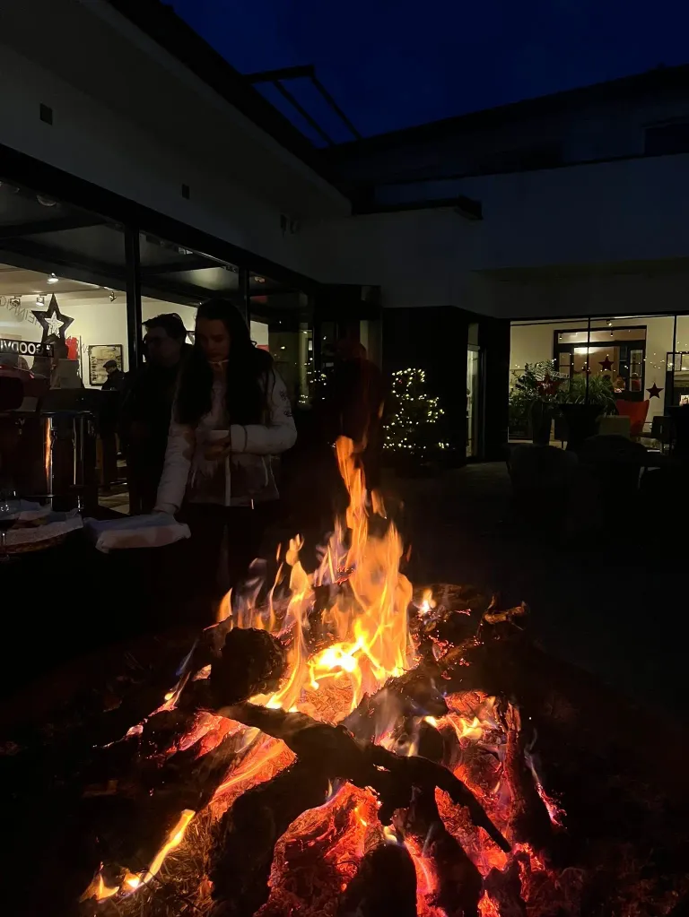 Eine Frau steht nachts vor einem Lagerfeuer mit Leuten um sie herum und genießt die Wärme und Atmosphäre. Das Gebiet ist mit Weihnachtslichtern dekoriert.