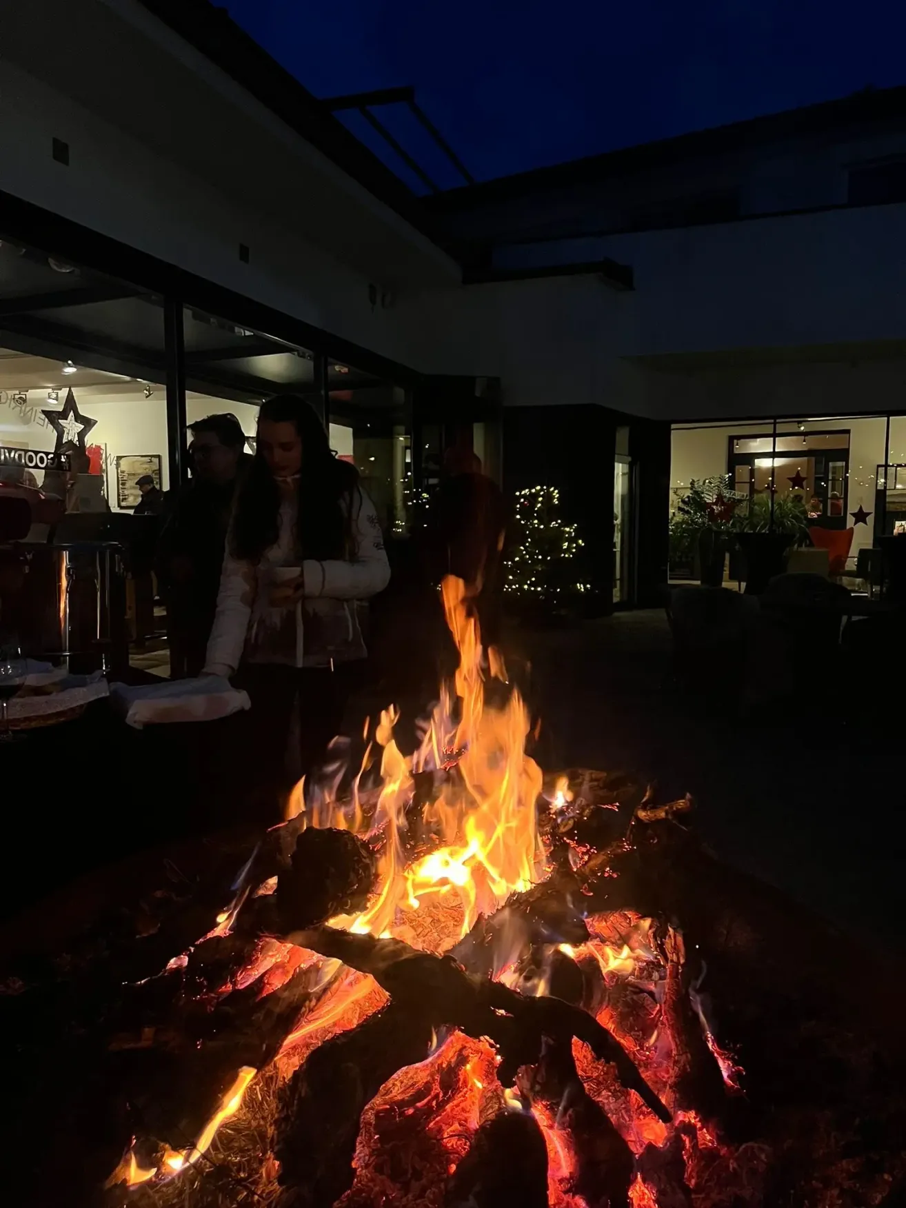 Eine Frau steht nachts vor einem Lagerfeuer mit Leuten um sie herum und genießt die Wärme und Atmosphäre. Das Gebiet ist mit Weihnachtslichtern dekoriert.