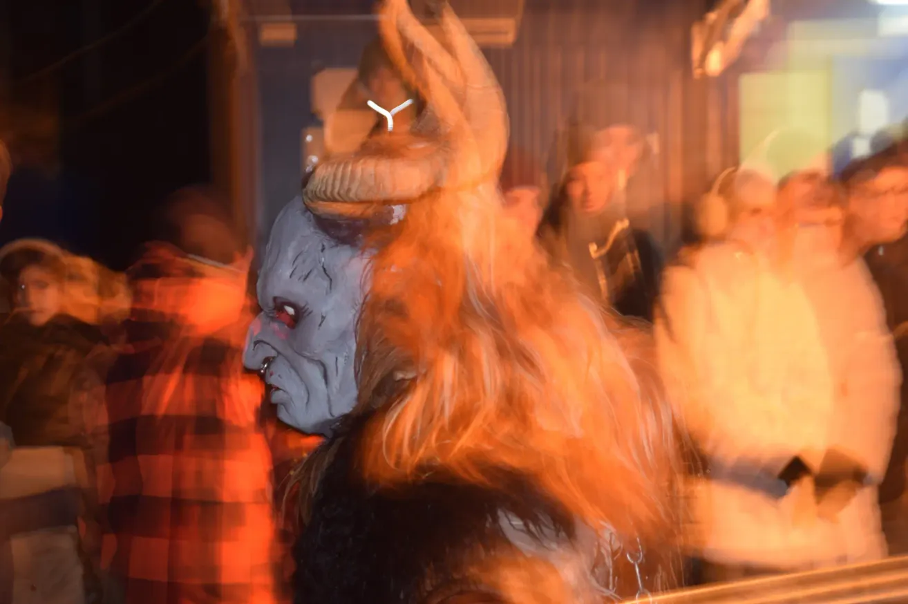 Eine Person mit einer langen, feuerroten, gehörnten Perücke und einer Maske steht in einem schwach beleuchteten Raum, mit anderen Menschen im Hintergrund.