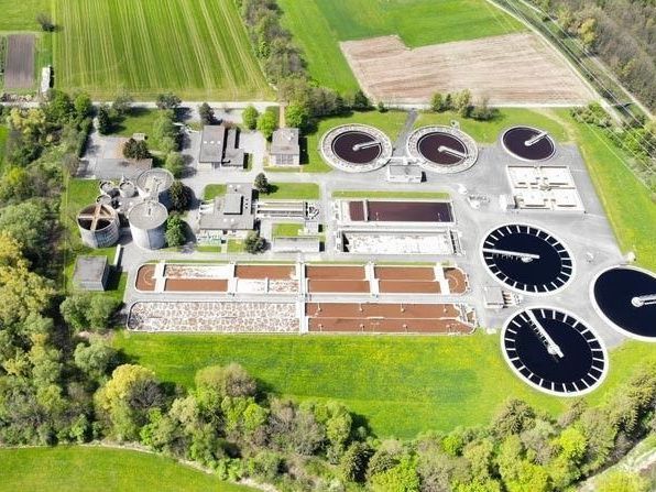 Ein Luftbild einer Abwasserbehandlungsanlage, umgeben von grünen Feldern und Bäumen. Mehrere große runde Tanks sind sichtbar, die Wasseraufbereitungsprozesse anzeigen. Gebäude und andere Strukturen sind ebenfalls vorhanden.