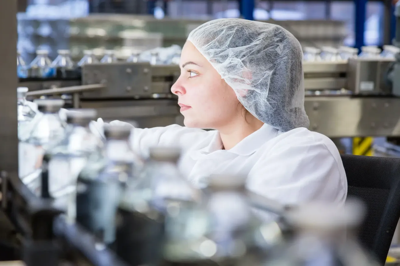 Eine Frau in weißer Laborkittel und Haarnetz arbeitet in einer Fabrik, möglicherweise inspiziert oder verpackt sie Flaschen auf einem Förderband.
