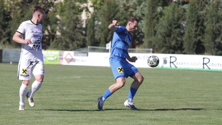 Ein Fußballspieler in einem blauen Trikot tritt einen Fußball auf einem Rasenfeld. Im Hintergrund sind Bäume und ein Zaun zu sehen.