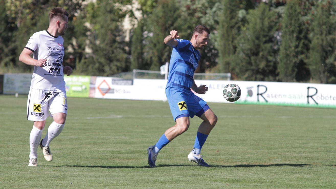 Ein Fußballspieler in einem blauen Trikot tritt einen Fußball auf einem Rasenfeld. Im Hintergrund sind Bäume und ein Zaun zu sehen.