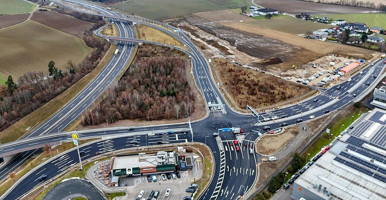 Eine Luftaufnahme einer belebten Autobahnkreuzung mit Autos, Lastwagen und einem McDonald's-Restaurant. Die Straße hat mehrere Spuren und einen Kreisverkehr. Im Hintergrund befinden sich Gebäude und Bäume.