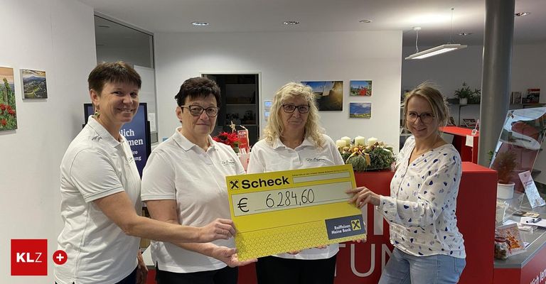 Vier Frauen halten einen großen gelben Scheck mit einem Betrag von 6.284,60 Euro. Sie stehen in einem Büro mit Weihnachtsdekoration und einem Schild für Raiffeisen Meine Bank.