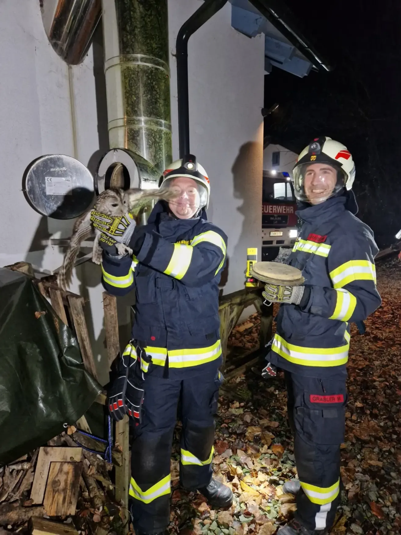 Zwei Feuerwehrleute stehen vor einem Haus, einer hält eine kleine Eule und der andere hält ein rundes Objekt, möglicherweise ein Feuerlöscherdeckel.