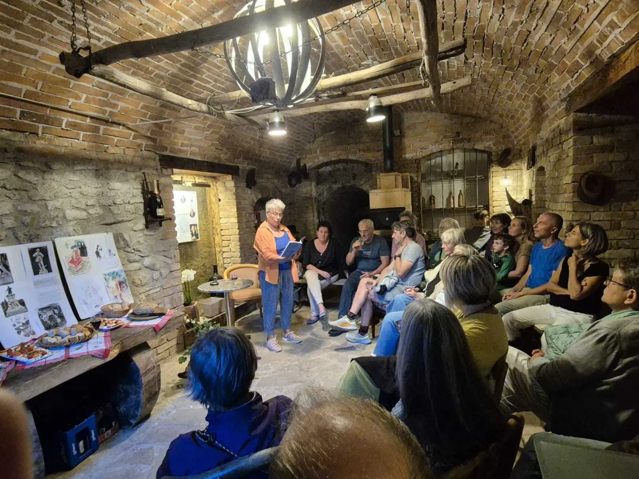 Eine Frau steht in einem Keller mit einer Ziegelwand und liest ein Buch. Menschen sitzen um sie herum und lauschen. Ein Tisch hält eine Blume, einen Teller mit Essen und eine Flasche. Eine Kronleuchter hängt von der Decke.