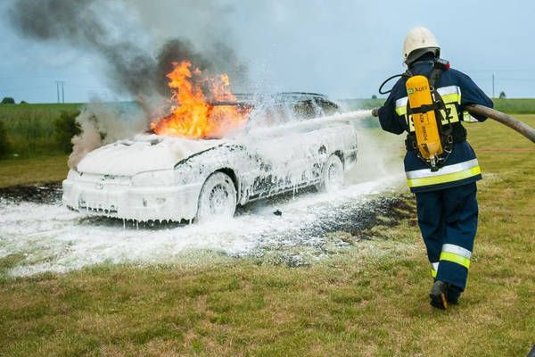Ein Feuerwehrmann löscht ein brennendes Auto mit einem Schlauch, das Fahrzeug ist mit Schaum bedeckt und Flammen sind sichtbar.