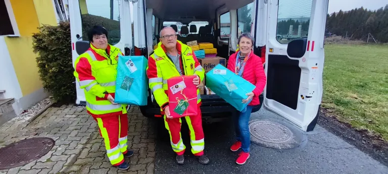 Drei Personen in Sicherheitsanzügen halten eingewickelte Geschenke in der Nähe eines offenen Lieferwagens mit vielen weiteren Geschenken im Inneren. Sie stehen auf einem gepflasterten Bürgersteig.