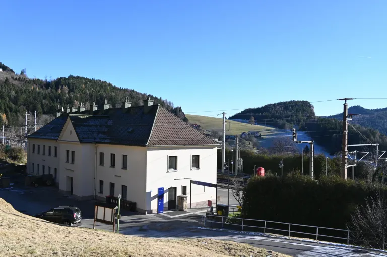 Ein weißes Gebäude mit braunem Dach steht in einem ländlichen Gebiet. Mehrere Fenster und eine blaue Tür sind sichtbar. Bäume und Berge im Hintergrund. Ein Schild steht in der Nähe des Eingangs.