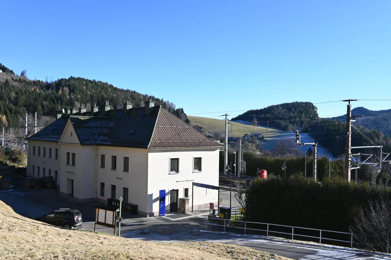 Ein weißes Gebäude mit braunem Dach steht in einem ländlichen Gebiet. Mehrere Fenster und eine blaue Tür sind sichtbar. Bäume und Berge im Hintergrund. Ein Schild steht in der Nähe des Eingangs.