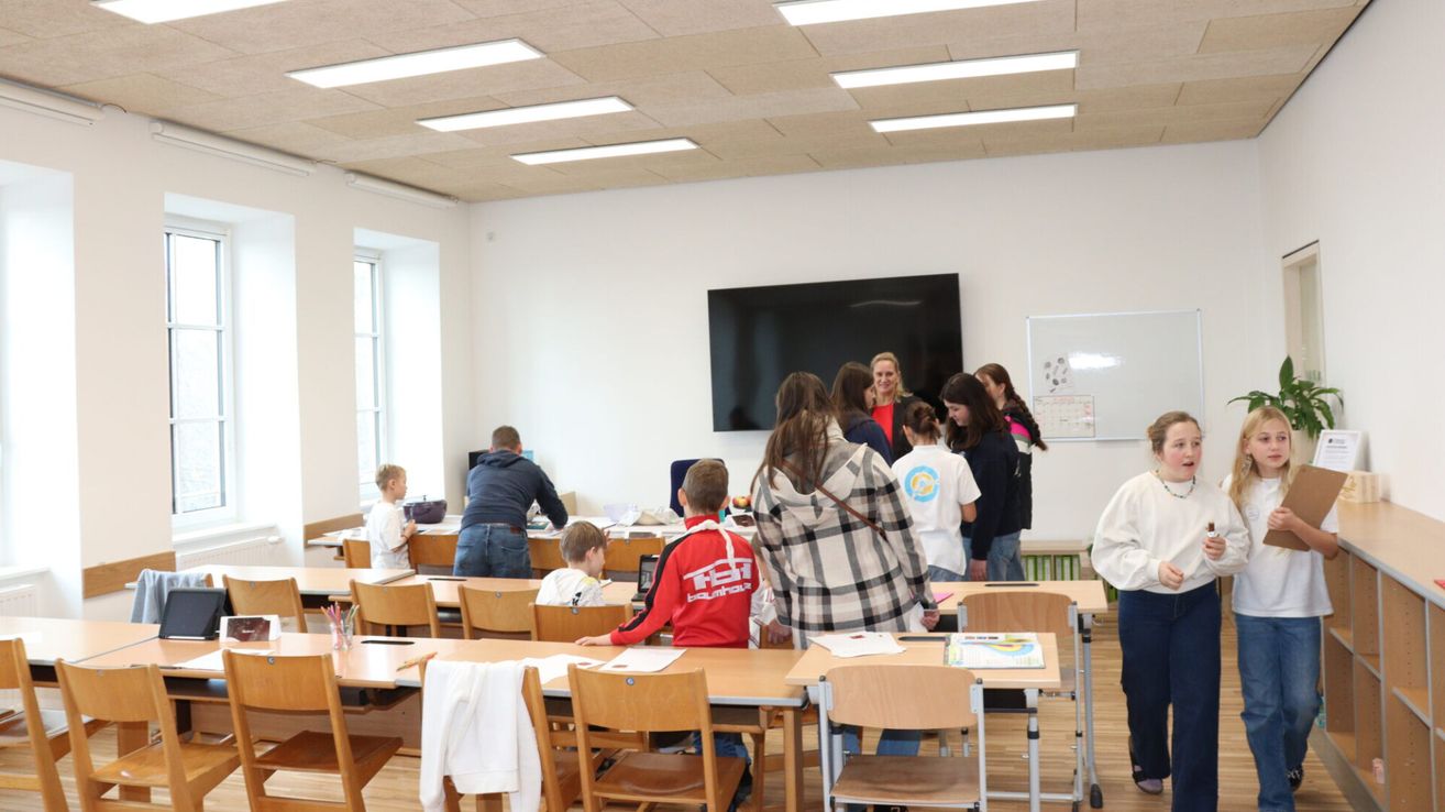 Ein Klassenzimmer mit Schülern und einer Lehrerin. Ein großer Fernseher ist an der Wand montiert und eine Tafel befindet sich rechts. Einige Schüler stehen in der Nähe der Lehrerin, während andere auf Stühlen vor Tischen sitzen.