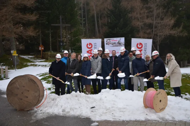 Eine Gruppe von Menschen in Helmen steht im Schnee, hält Schaufeln und posiert für ein Foto. Sie stehen vor Bannern mit den Worten BAG und PROGRESS. Dahinter befinden sich Bäume und ein Schild.