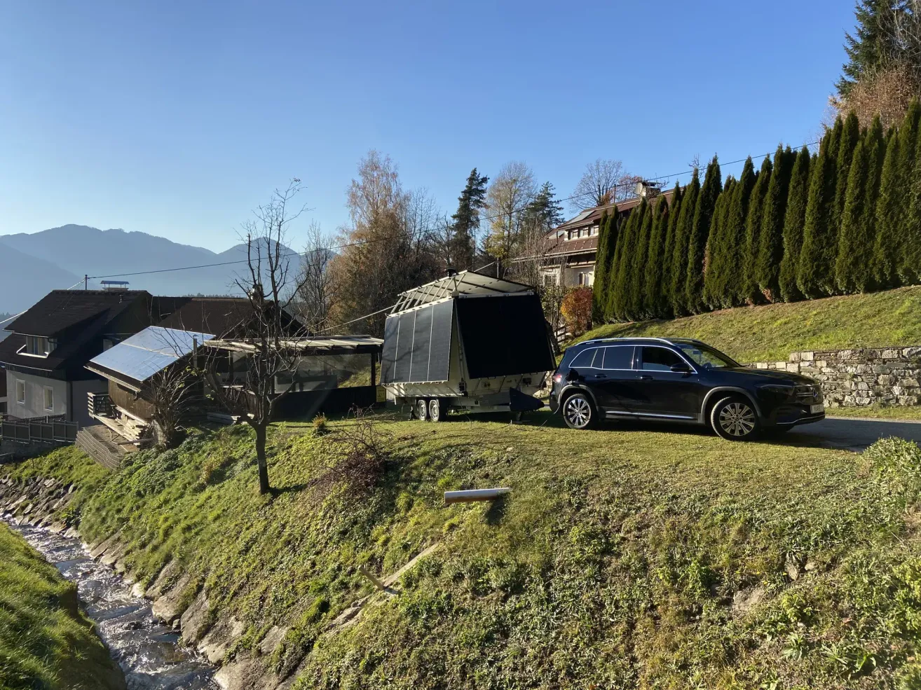 Ein schwarzer SUV ist auf einem Grashang neben einem Anhänger mit Solarpanelen geparkt. In der Nähe befindet sich ein kleines Haus mit einem Solarpanel auf dem Dach. Bäume und eine Hecke sind am Hang, mit einem Gebäude und Bergen in der Ferne.