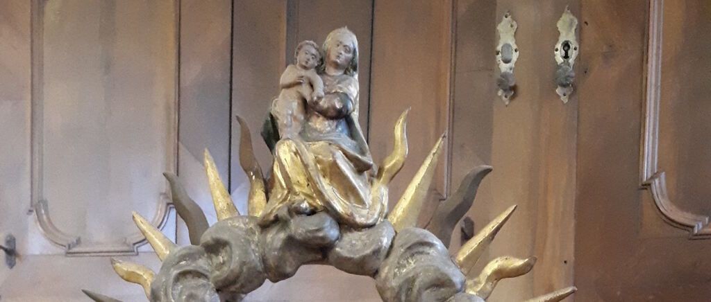 Eine goldene Statue von Maria und Jesus sitzt auf einer Wolke, geschmückt mit Lichtstrahlen. Die Statue steht an einer braunen Wand mit zwei Türschlössern.
