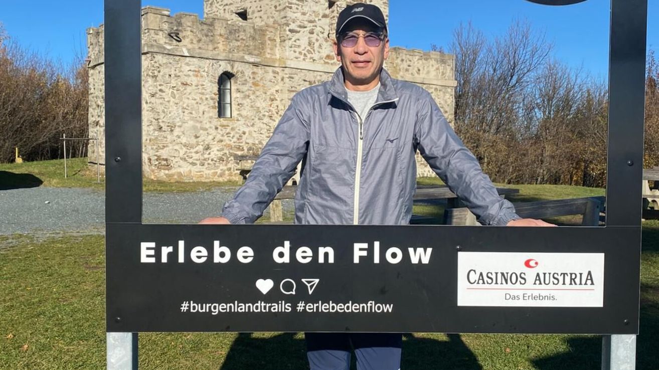 Ein Mann in einer grauen Jacke steht vor einem Steingebäude und hält ein Schild mit der Aufschrift 'Erlebe den Flow' mit dem Casino-Logo und Hashtags.