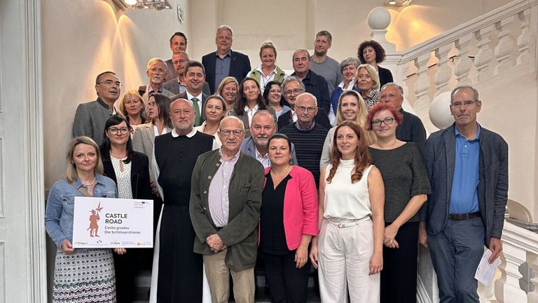 Eine Gruppe von Menschen, darunter Erwachsene und ältere Menschen, posiert für ein Foto auf einer Treppe. Sie lächeln und scheinen glücklich zu sein. Einige von ihnen tragen Brillen und einer hält ein weißes Papier mit Text.