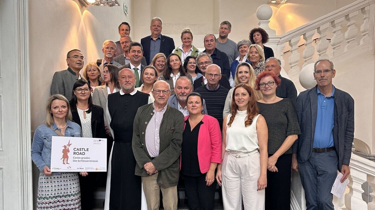 Eine Gruppe von Menschen, darunter Erwachsene und ältere Menschen, posiert für ein Foto auf einer Treppe. Sie lächeln und scheinen glücklich zu sein. Einige von ihnen tragen Brillen und einer hält ein weißes Papier mit Text.