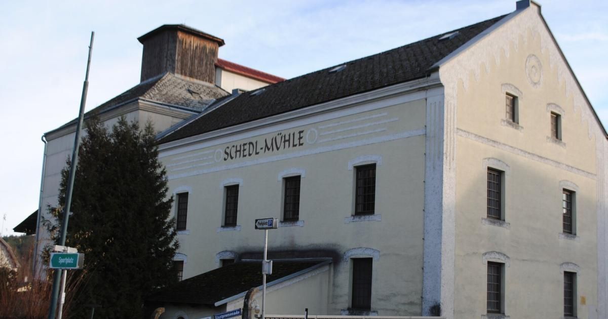 Ein altes Gebäude mit einem Schornstein und dem Schild Schedl-Mühle über dem Eingang. Mehrere Fenster und ein Straßenschild mit der Aufschrift Parken P.