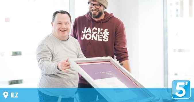 Zwei Männer lächeln und posieren mit einem gerahmten Bild. Einer trägt ein graues Langarmshirt, der andere eine dunkelrote Jack & Jones-Kapuzenjacke. Ein Wasserzeichen mit der Nummer 5 in der Ecke.