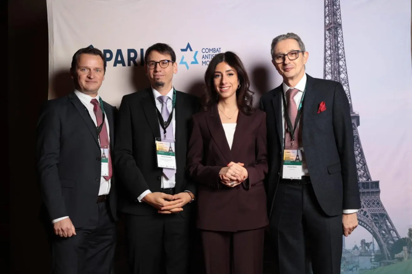 Vier Personen stehen eng beieinander und tragen formelle Kleidung. Der Mann rechts trägt eine rote Anstecknadel. Hinter ihnen befindet sich ein Banner mit der Aufschrift 'COMBAT ANTISEMITISM MOVEMENT'.