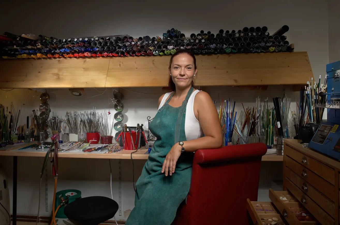 Eine Frau in einem grünen Apron sitzt auf einem roten Stuhl in einem Kunstatelier, umgeben von Kunstmaterialien und Werkzeugen auf Regalen und einem Schreibtisch.
