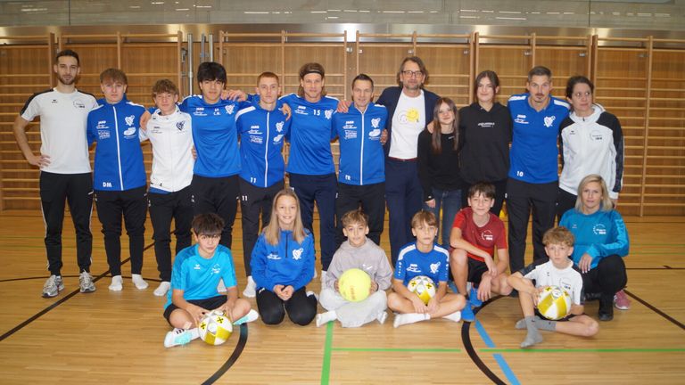 Ein Sportteam posiert für ein Foto in einer Turnhalle. Sie tragen passende blaue Trikots und einige halten Sportbälle. Zwei Personen in der Mitte tragen eine Sonnenbrille und ein Lächeln auf ihrem Shirt.