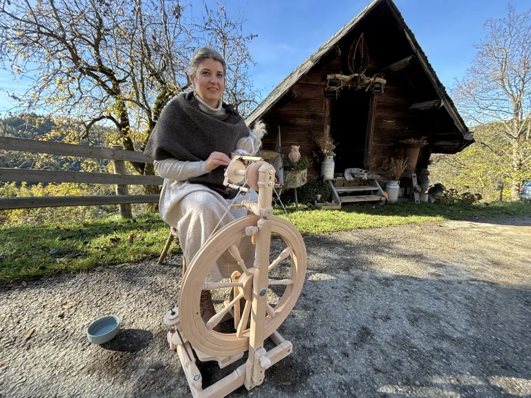 Eine Frau sitzt auf einem Holzstuhl und bedient eine Spinnmaschine, mit einem Holzhaus und einem Holzzaun im Hintergrund.