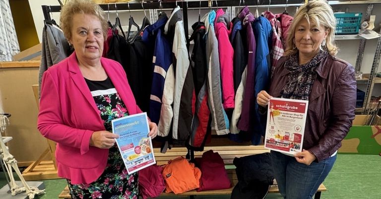 Zwei Frauen stehen vor einem Kleiderständer mit bunten Jacken. Sie halten Flyer in den Händen und lächeln für die Kamera.