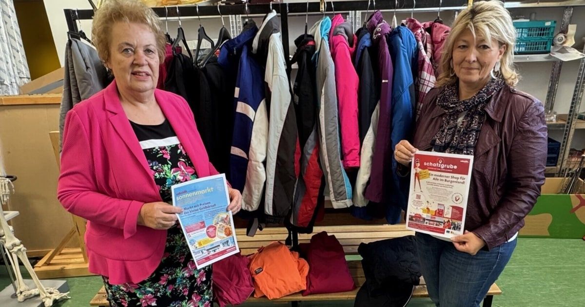 Zwei Frauen stehen vor einem Kleiderständer mit bunten Jacken. Sie halten Flyer in den Händen und lächeln für die Kamera.