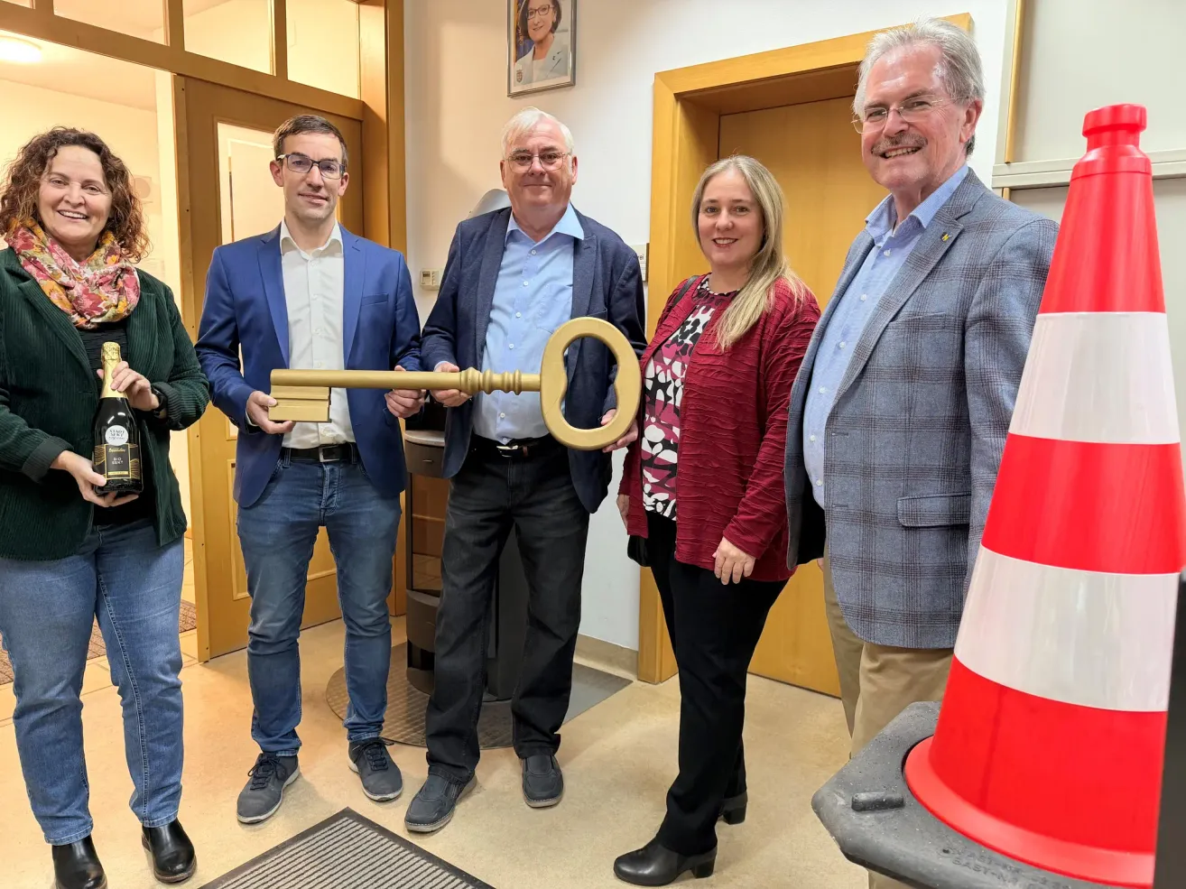 Fünf Erwachsene stehen in einem Raum. Zwei Männer und eine Frau halten einen goldenen Schlüssel. Sie lächeln. Hinter ihnen steht ein Verkehrskegel.