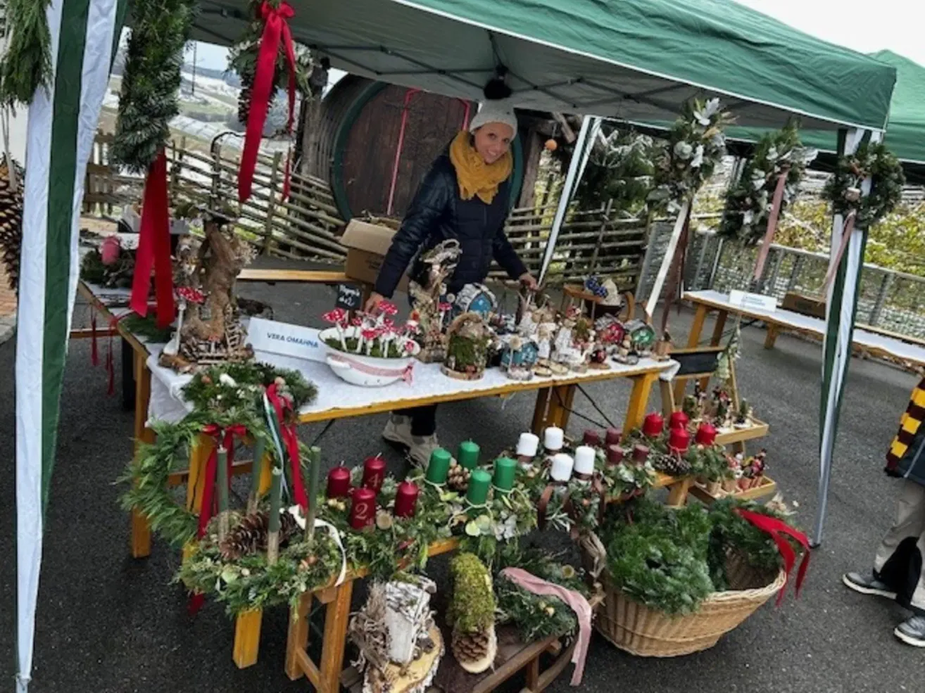 Ein Weihnachtsmarkt-Display unter einem grünen Zelt mit verschiedenen Ornamenten, Kränzen und Figuren. Eine Frau in Mütze und Schal steht hinter dem Tisch.