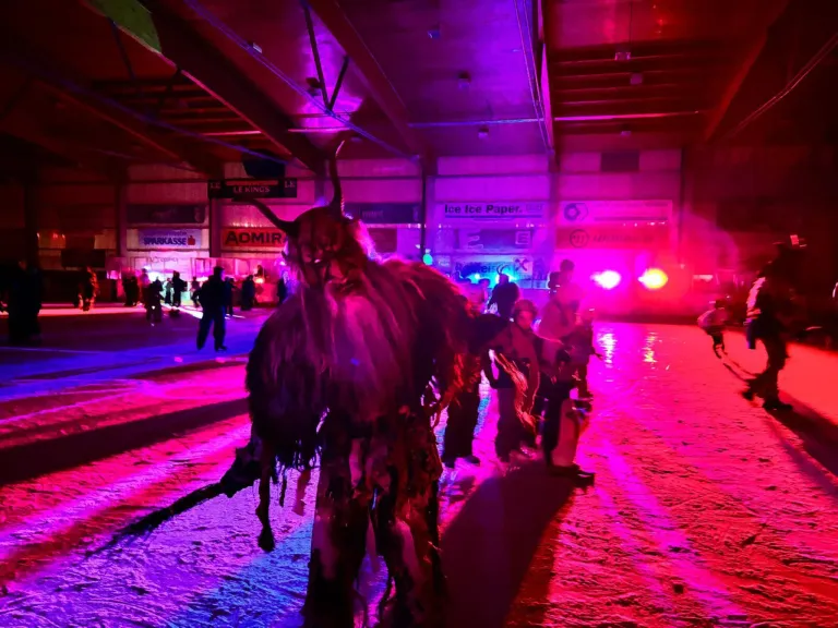 Eine Person in einem Hörnerkostüm steht in einem schwach beleuchteten Bereich mit lila Beleuchtung, möglicherweise für eine Halloween-Veranstaltung. In der Nähe stehen und gehen Menschen.